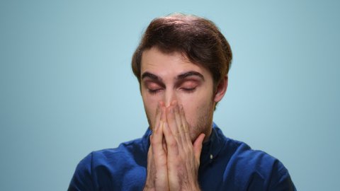 Sick man sneezing in studio. Close up ill guy sneezing on blue background. Portrait of ill man sneezing from allergy in slow motion. Male model holding hands on face. Sick student posing at camera
