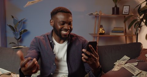 Happy afro american man holding smartphone and making successful deal while working from home. Young guy winning big money and rejoicing. Money falling from above.