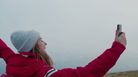 Free Young Female Hiker in Red Jacket Making Video Call Waving Hello to Friends and Sending Air Kisses from Top of Mountain. Happy Smiling Girl Recording Selfie Video Using Smartphone. Slow Motion