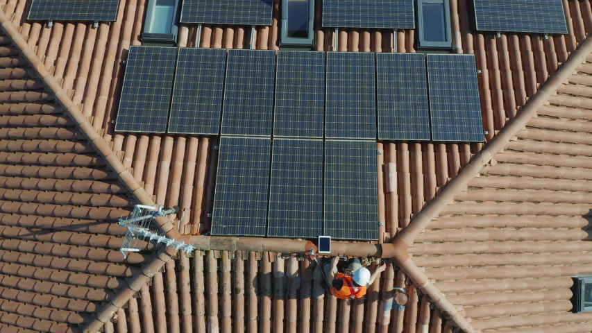 Aerial zenithal shot of technician with hard hat and safety equipment installing photovoltaic solar panel system installed on home domestic roof top, urban landscape. eco responsible business concept | Shutterstock HD Video #1051269976