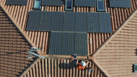 Aerial zenithal shot of technician with hard hat and safety equipment installing photovoltaic solar panel system installed on home domestic roof top, urban landscape. eco responsible business concept
