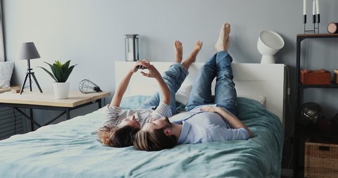Carefree young adult couple enjoying relaxing lying in bed using modern smartphone technology. Man and woman making video call, playing mobile games, watching video, taking selfie in bedroom together.