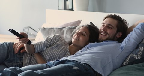 Happy relaxed young family couple lying in bed holding remote control watching tv together. Smiling millennial man and woman lounging embracing talking enjoying television in cozy bedroom at home.