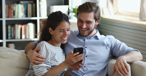 Happy millennial couple holding smartphone, talking, playing mobile game, laughing sitting on sofa. Smiling man and woman customers doing shopping on cellphone using easy app order delivery from home.
