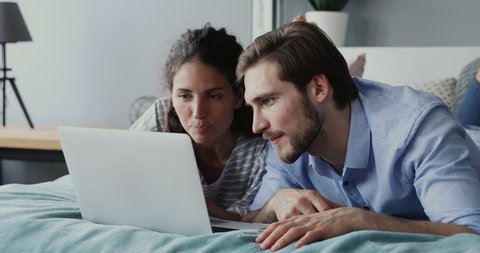Happy millennial man and woman using laptop computer at home watching online show lying in bed together. Young adult couple looking at notebook screen making video call chatting in bedroom at home.