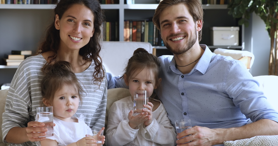 Healthy family young adult parents with small kids drinking clean fresh pure water together looking at camera at home. Happy couple and cute little children health care hydration concept. Portrait Royalty-Free Stock Footage #1051271872