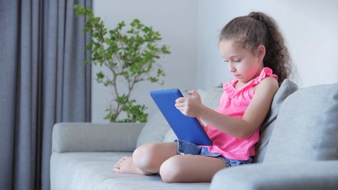 Curious cute kid little girl using digital tablet technology device included in studies sitting on sofa at home alone. Small child baby hold pad computer play games at home.Children tech addiction