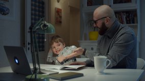 Young 30s father gives high five to son while making online homework at home. Bearded man and young boy studying online with laptop. 