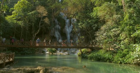 Aerial shot of tourists exploring beautiful waterfall amidst trees in rainforest, drone flying backward over people on footbridge during sunny day - Luang Prabang, Laos
