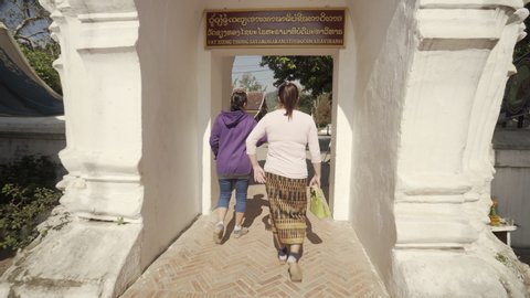 Women walking through white archway at historic famous temple on sunny day - Luang Prabang, Laos