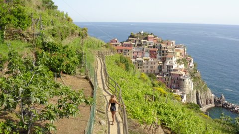 Aerial shot of woman walking on footpath by plants near city, drone ascending from young tourist on mountain near sea - Cinque Terre, Italy