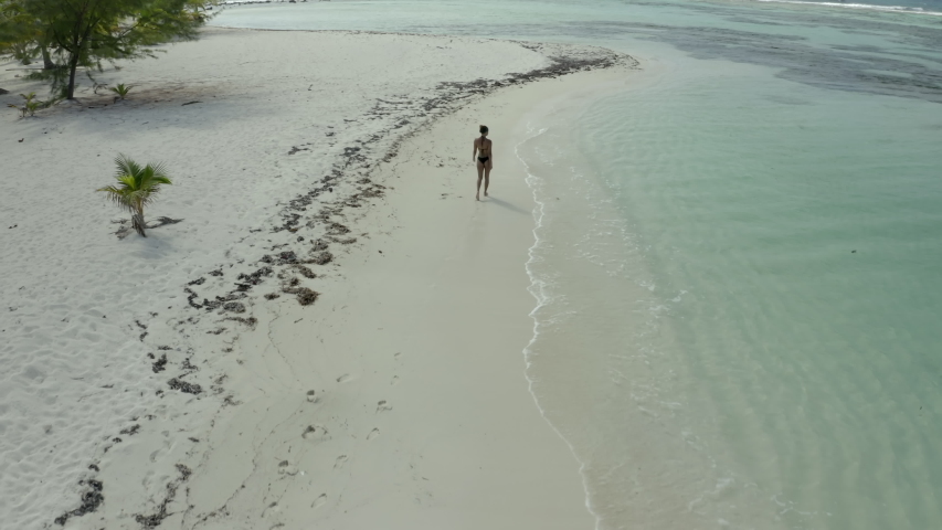 Aerial: Drone moving towards woman in bikini walking on sea shore, she in on vacation at beach - Coco Plum Cay, Belize