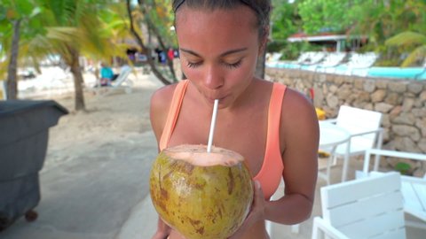 Slow motion of beautiful woman drinking fresh from coconut at beach, young female tourist wearing bikini by furniture - Montego Bay, Jamaica