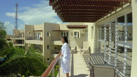Aerial: Young woman removing bathrobe in balcony at hotel, coconut palm trees by buildings near sea against sky on sunny day - Great Blue Hole, Belize