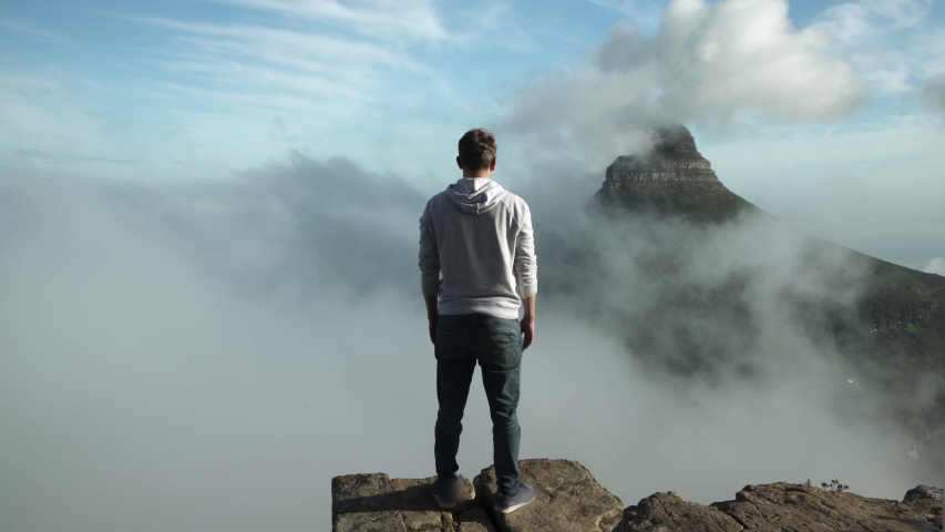Time lapse shot from the back of a man standing on a cliff over amazing clouds moving around a mountain - Cape Town, South Africa