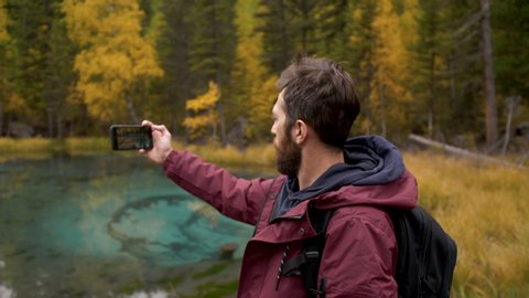 30s Forest Hiker Photographer Man in Woods Shooting Lake View Back Shot Concept. Contemporary Travel Guy Videographer Standing in Nature Scenic and Natural Filming via Panoramic Hold Smartphone Camera