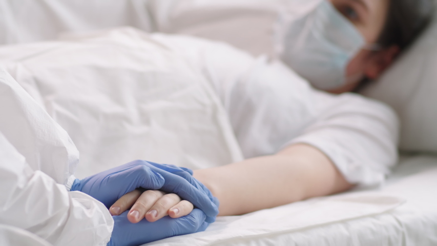 Selective focus shot of female coronavirus patient in protective face mask lying on couch in hospital ward while nurse in sterile gloves and suit stroking her hand, supporting and soothing her | Shutterstock HD Video #1051329520