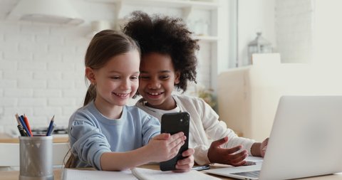Two happy cute diverse ethnicity children using smartphone app for remote mobile education having fun playing or studying together at home. Small african and caucasian girls having fun using cell.