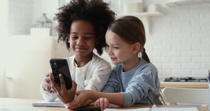 Cute mixed race kids girls friends or sisters playing with smart phone together holding gadget obsessed with social media, learning in app at home. Diverse children mobile technology addiction concept