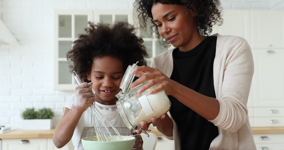 African mom and small kid daughter cooking together making dough in bowl in kitchen. Happy mixed race family prepare cake mix ingredients for pie having fun baking pastry on holiday morning at home. Royalty-Free Stock Footage #1051329874