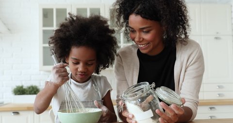 African mom and small kid daughter cooking together making dough in bowl in kitchen. Happy mixed race family prepare cake mix ingredients for pie having fun baking pastry on holiday morning at home.
