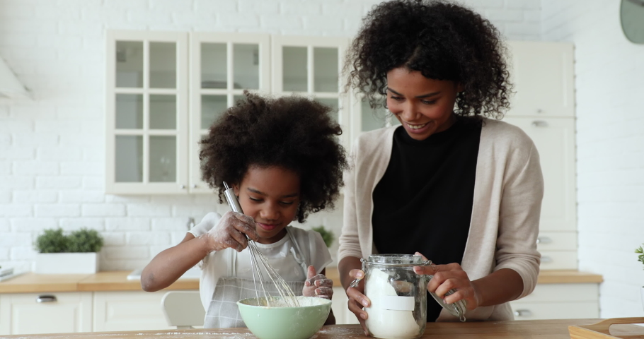 Happy afro american young adult mum and little cute child daughter whisking dough in bowl making holiday cake or pancakes at home. Mixed race family baking pastry mixing flour to recipe in kitchen. Royalty-Free Stock Footage #1051329877