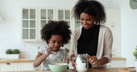 Happy afro american young adult mum and little cute child daughter whisking dough in bowl making holiday cake or pancakes at home. Mixed race family baking pastry mixing flour to recipe in kitchen.