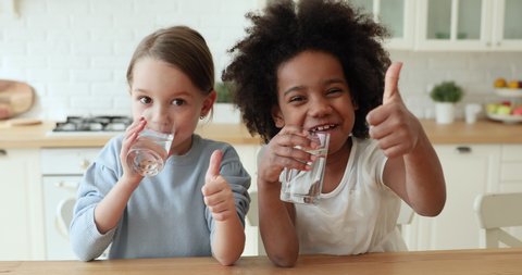 Two funny adorable mixed race children girls holding glasses drinking tasty clean pure mineral water showing thumbs up looking at camera, recommend daily children healthcare morning hydration concept.