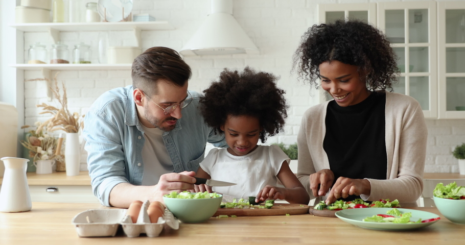 Happy mixed race family of three young parents and afro american kid daughter enjoy cooking together. Multi ethnic mum and dad teaching cute child learning making healthy vegetable salad in kitchen. Royalty-Free Stock Footage #1051329892
