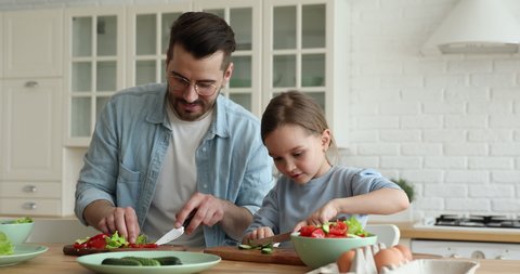 Young father teaching cute kid daughter cutting fresh vegetable salad in kitchen. Single adult parent dad explaining small child girl learning cooking healthy food prepare vegan meal together at home.