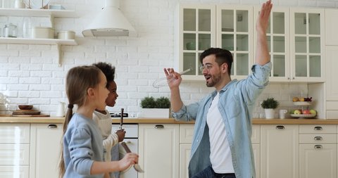 Happy biracial family young adult father and two cute mixed race kids having fun dancing and singing in kitchen. Funny dad holding kitchenware microphone playing with multiracial children at home.
