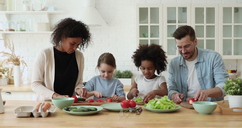 Happy mixed race parents and kids daughters enjoy cooking together. Multiracial family african mom, caucasian dad teaching cute diverse children cutting vegetable salad prepare healthy meal together.