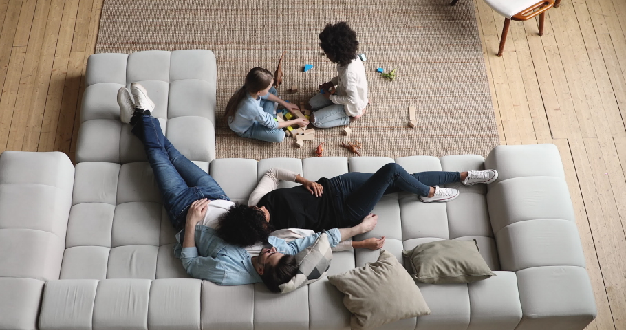 Mixed ethnicity parents relaxing on comfortable couch while cute diverse kids daughters playing on floor at home. Interracial family with two children spending time together in living room. Top view | Shutterstock HD Video #1051329919