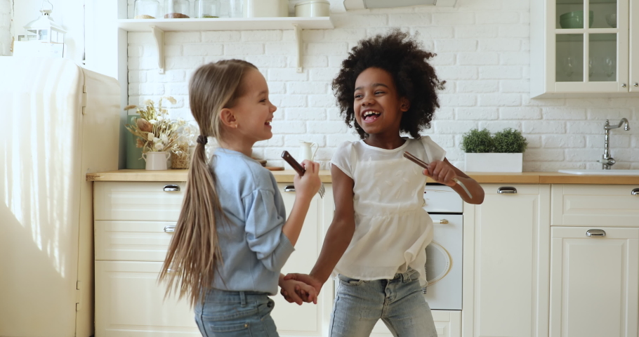 Happy cute african and caucasian small girls stepsisters or friends having fun playing together in kitchen. Two funny mixed race children dancing, jumping, singing in kitchenware microphones at home. | Shutterstock HD Video #1051329922