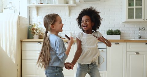 Happy cute african and caucasian small girls stepsisters or friends having fun playing together in kitchen. Two funny mixed race children dancing, jumping, singing in kitchenware microphones at home.