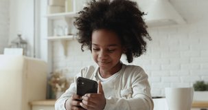 Cute african kid girl holding smart phone enjoying using mobile apps, playing games at home. Small mixed race child learning in cellphone, watching video, having fun with mobile technology concept.