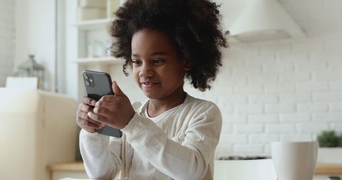 Cute african kid girl holding smart phone enjoying using mobile apps, playing games at home. Small mixed race child learning in cellphone, watching video, having fun with mobile technology concept.
