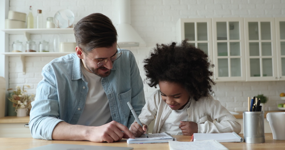 Mixed ethnicity family caucasian stepfather and african school child daughter study together sit at kitchen table. Male tutor helping ethnic kid girl explaining learning math doing homework at home. | Shutterstock HD Video #1051329967