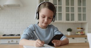 Cute child girl wearing headphones studying online in mobile app by video call with remote teacher, tutor. School kid holding phone doing homework using application, watching class learning at home.