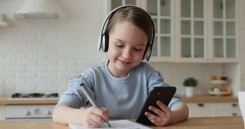 Cute child girl wearing headphones studying online in mobile app by video call with remote teacher, tutor. School kid holding phone doing homework using application, watching class learning at home.