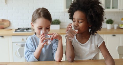 Healthy cute mixed race kids girls drinking water at home. Adorable african and caucasian children hydrating thirst to prevent dehydration sit at kitchen table. Children health care nutrition concept.