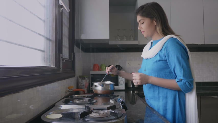 A beautiful attractive Indian woman or female housewife or lady in salwar kameez or traditional costumes or cloths dress cooking food on a burner gas stove in an indoor kitchen.  Royalty-Free Stock Footage #1051332043