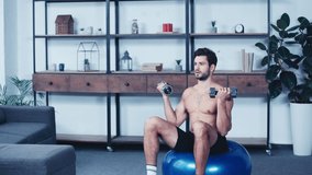 young shirtless sportsman sitting on fitness ball and training with dumbbells