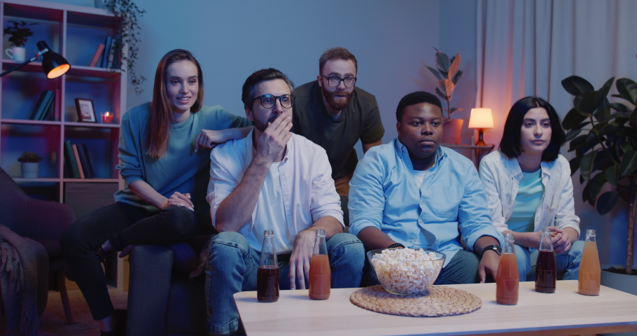 Group of young friends watching sport game with serious face while sitting on sofa. Concentrated people watching competition on TV in front of table with drinks and popcorn on it. | Shutterstock HD Video #1051341766