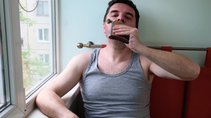 Pensive 40 years old man sitting at home near a window, drinking alchole from a metal flask,. Quarantine, self isolation, stay at home concept.