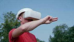 Man in a virtual reality helmet controls gestures. The guy in the park in a red T-shirt on a background of blue sky and green trees looks VR glasses. Place for graphics. 4K footage