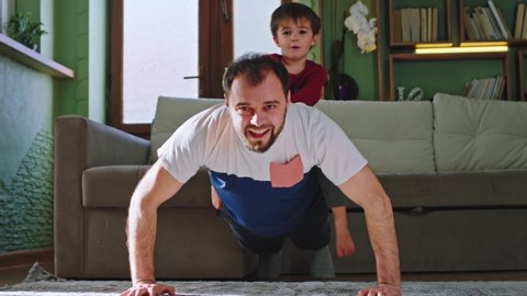 Funny little boy with his dad doing sport at home in living room at sunset dad doing hard push-ups with small boy on his back. Shot on ARRI Alexa Mini