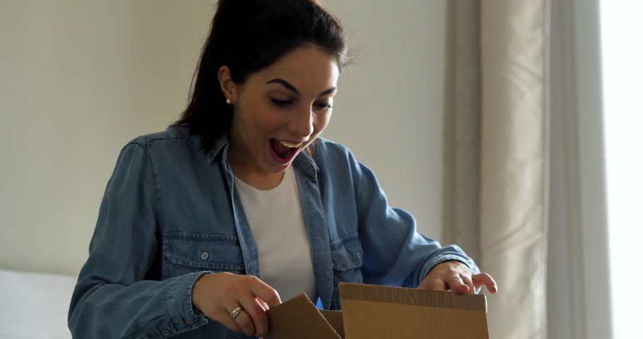 An young woman is excited while opening a package of a parcel delivered by mailman in her home. Concept: purchase, delivery, e-commerce, online shopping, emotions, client satisfaction, surprise