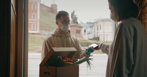 postman or delivery asian man carry small box deliver to young woman customer at home contactless nfc terminal payment. Man wearing mask prevent covid or coranavirus quarantine pandemic. 