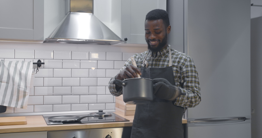 Portrait of cheerful young afro-american guy in apron preparing food in pan stirring with wooden spoon making meal for dinner in modern small kitchen Royalty-Free Stock Footage #1051388041